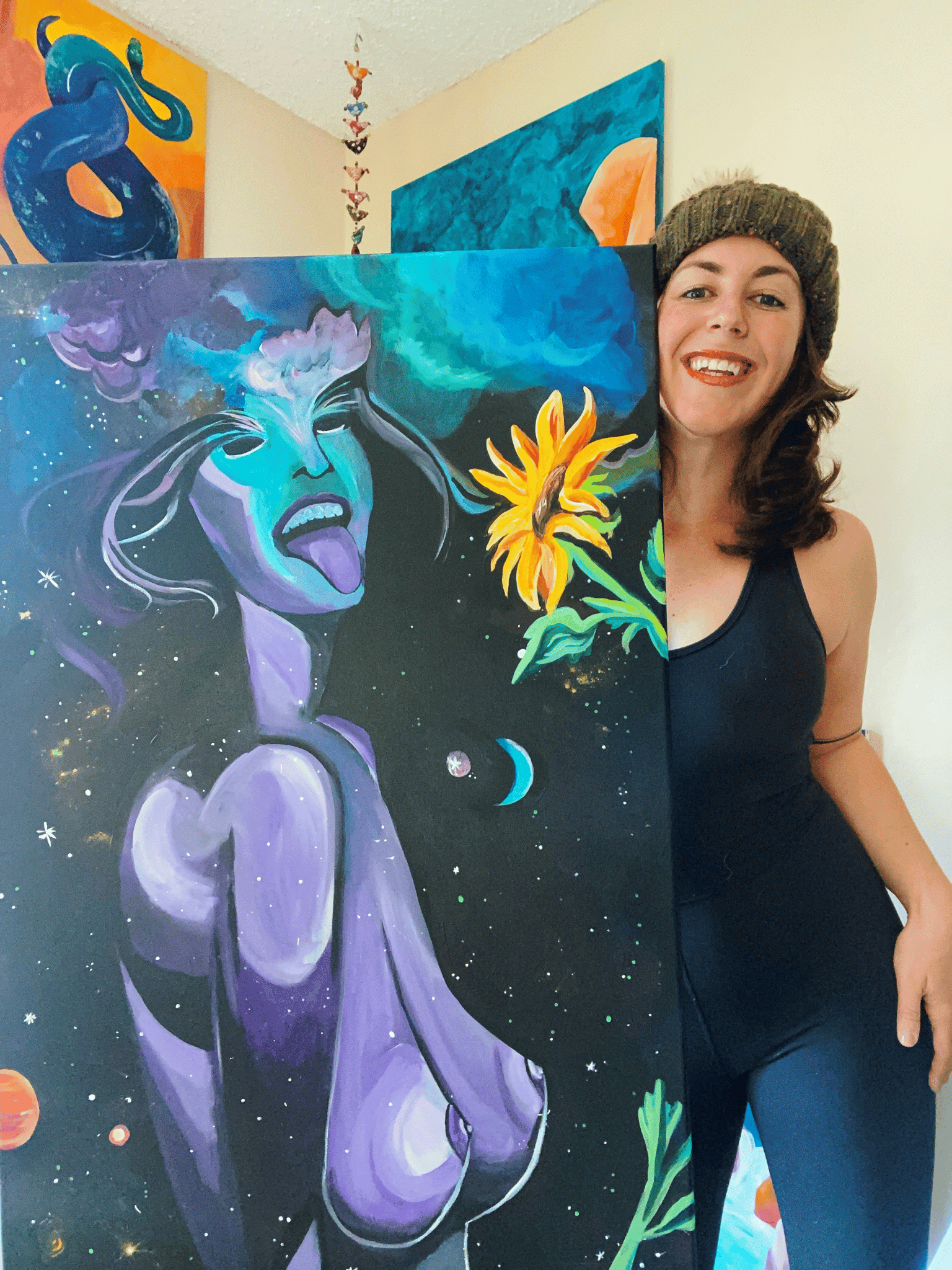 Untitled design (12) image of Kate DeCoste holding up her painting of a nake woman, her tongue sticking out in space with a sunflower